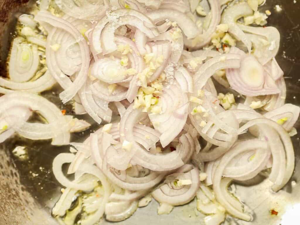 Close-up of thinly sliced shallots and minced garlic sizzling in a pan, creating a base for cooking. The ingredients are lightly browning and releasing a savory aroma.